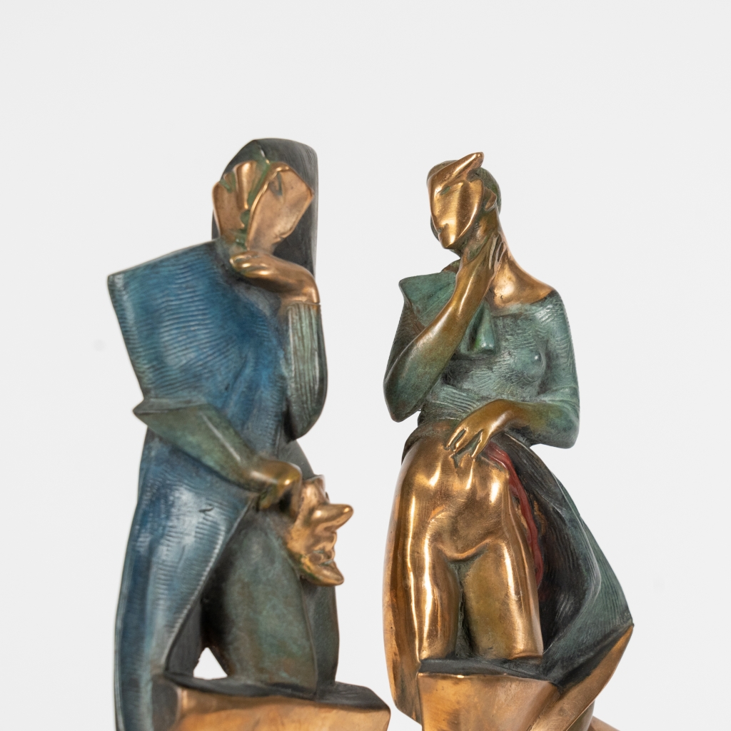 Plan sur le couple de Mascarade III d'Arsen Avetissian, sculpture contemporaine en bronze représentant deux personnages stylisés dominant deux grands masques se faisant face. Les patines dorées, vertes et bleutées renforcent le caractère symbolique de cette composition élégante.