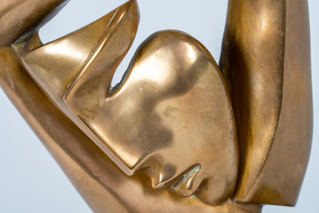 Zoom sur le visage de Un peu de soleil dans le froid d'Arsen Avetissian, sculpture contemporaine en bronze représentant un visage féminin stylisé encadré par deux bras levés dont les mains se rejoignent avec grâce. Les lignes fluides et la patine dorée créent une composition élégante, montée sur un socle en pierre.