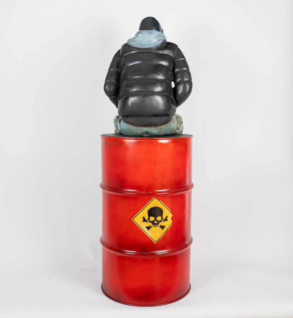Plan recto de l'Imposante sculpture en bronze grand format intitulée JULES, réalisée par Annabelle DEAIGUE suivant le processus de création à la cire perdue. Oeuvre représentant un jeune homme assis sur un baril de pétrol rouge.