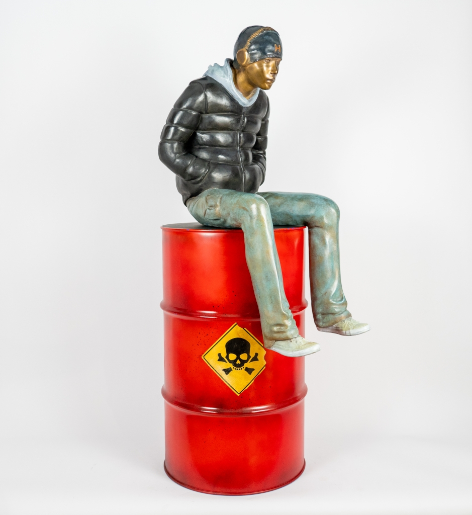 Plan de trois quarts de l'Imposante sculpture en bronze grand format intitulée JULES, réalisée par Annabelle DEAIGUE suivant le processus de création à la cire perdue. Oeuvre représentant un jeune homme assis sur un baril de pétrol rouge.