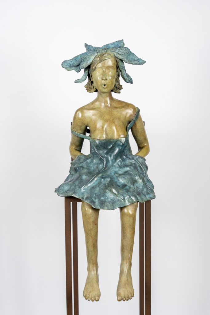Plan sur la sculpture originale en bronze de Cécile Cuvelier intitulée « Lolette », représentant une jeune femme assise sur un haut tabouret. Vêtue d'une robe aux mouvements fluides et coiffée d'un délicat ornement végétal, elle semble chanter ou souffler doucement, dans une posture pleine de grâce, de poésie et de légèreté.