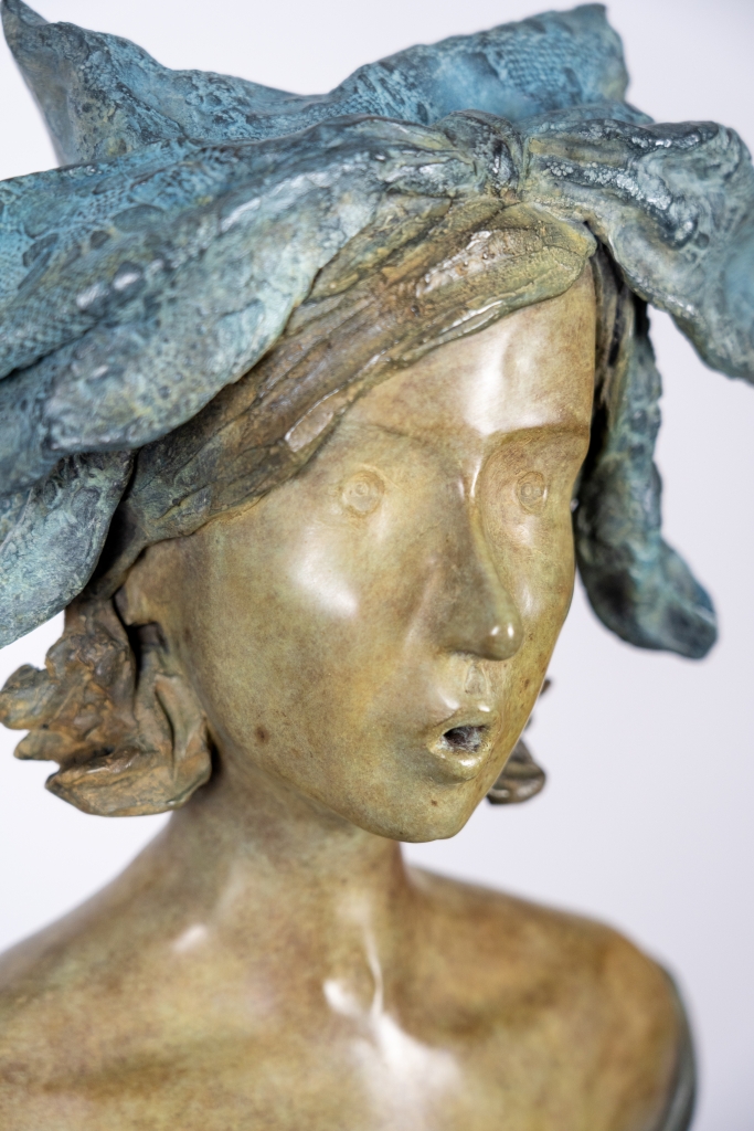 Zoom sur la sculpture originale en bronze de Cécile Cuvelier intitulée « Lolette », représentant une jeune femme assise sur un haut tabouret. Vêtue d'une robe aux mouvements fluides et coiffée d'un délicat ornement végétal, elle semble chanter ou souffler doucement, dans une posture pleine de grâce, de poésie et de légèreté.
